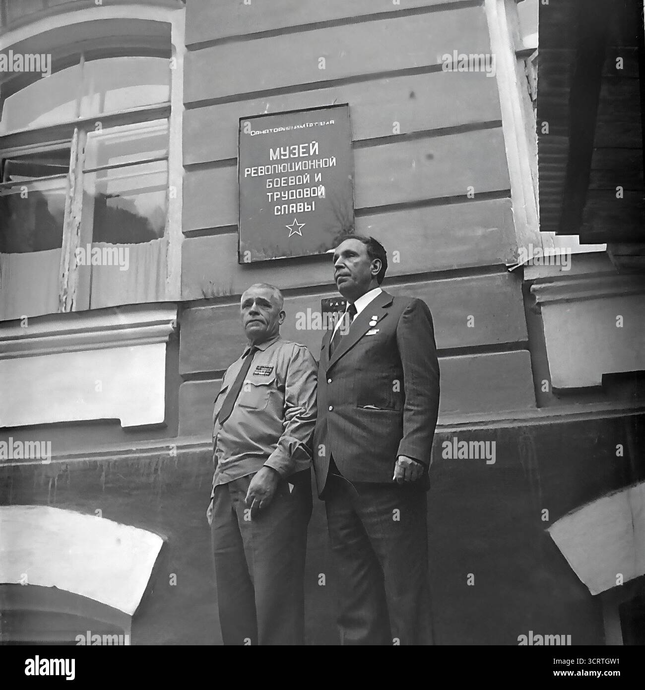 Un ritratto d'archivio formale in bianco e nero degli anni '1960, che mostra un veterano di guerra sovietico decorato e un funzionario del Partito Comunista in posa insieme a Sviatohirsk, RSS Ucraina. Sono orgogliosamente sotto il segno del locale "Museo della Gloria rivoluzionaria, militare e del lavoro". Questa fotografia è una rappresentazione simbolica classica dei pilastri della società sovietica: L'eroico veterano che incarna i sacrifici passati della nazione, e l'ufficiale che rappresenta il potere attuale dello stato, unito sotto la bandiera della storia istituzionalizzata Foto Stock