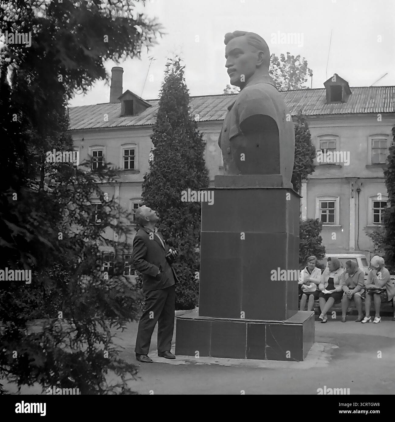 Un'autentica fotografia d'archivio in bianco e nero degli anni '1970 mostra un uomo sovietico in posa per una foto ricordo accanto a un grande monumento a Sviatohirsk, RSS Ucraina. L'uomo in tuta si erge orgogliosamente accanto al monumentale busto di una figura comunista. La scena, probabilmente ambientata sul terreno di un sanatorio o in un parco pubblico, è un tipico esempio di turismo dell'era sovietica, dove i visitatori spesso si fotografavano con punti di riferimento ideologici. Sullo sfondo, un gruppo di donne può essere visto riposare su una panchina Foto Stock