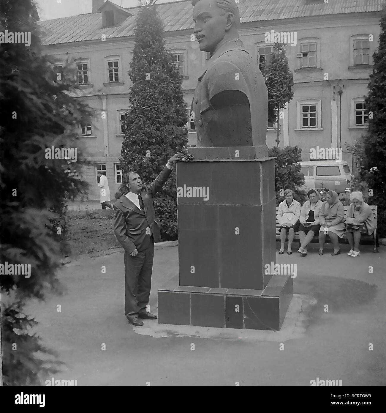 Un'autentica fotografia d'archivio in bianco e nero degli anni '1970 mostra un uomo sovietico in posa per una foto ricordo accanto a un grande monumento a Sviatohirsk, RSS Ucraina. L'uomo in tuta si erge orgogliosamente accanto al monumentale busto di una figura comunista. La scena, probabilmente ambientata sul terreno di un sanatorio o in un parco pubblico, è un tipico esempio di turismo dell'era sovietica, dove i visitatori spesso si fotografavano con punti di riferimento ideologici. Sullo sfondo, un gruppo di donne può essere visto riposare su una panchina Foto Stock