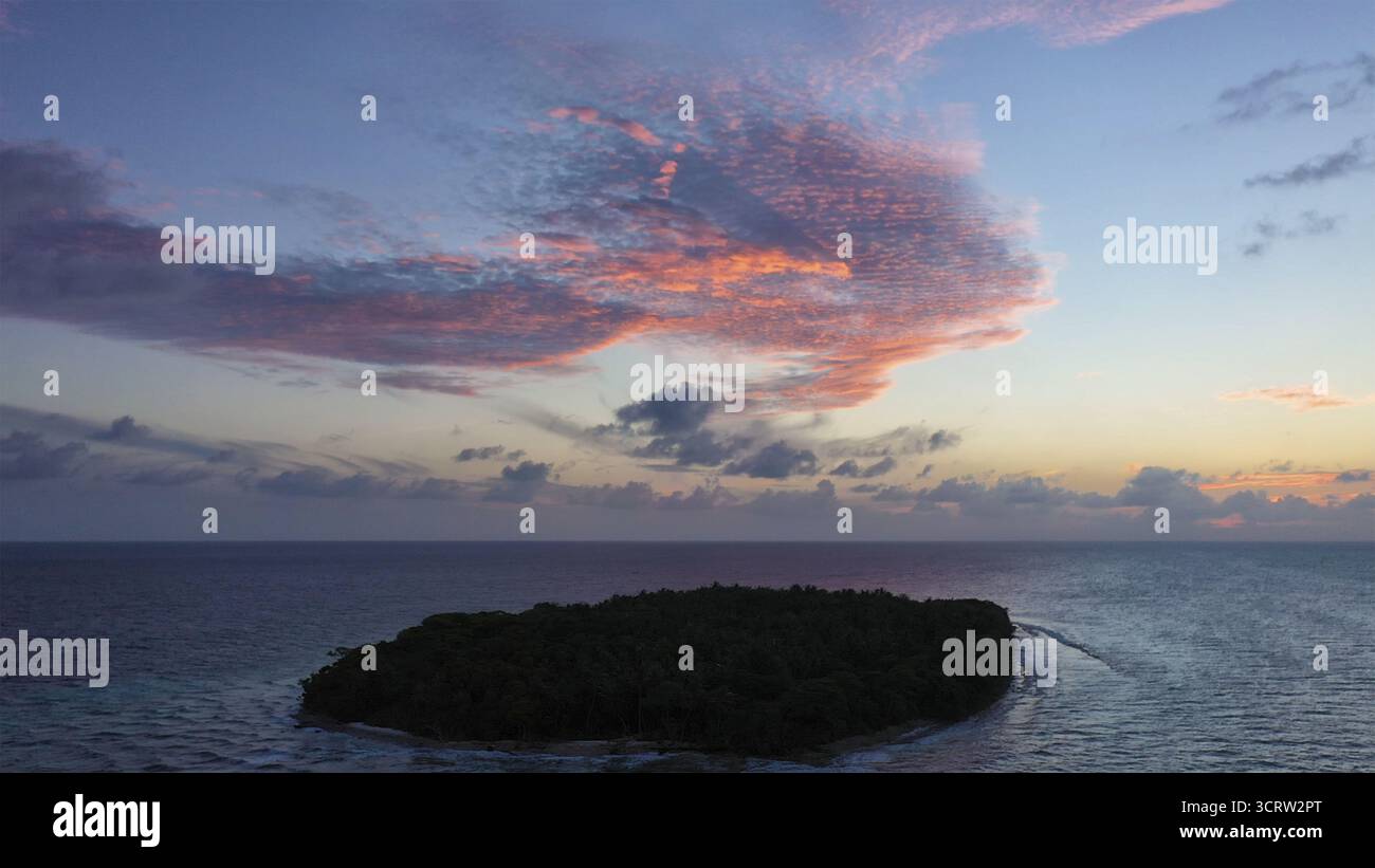 Vista aerea di un'isola verdeggiante e densamente boscosa circondata dal vasto e scuro oceano sotto un cielo dipinto con sfumature di rosa e blu, Maldive. Foto Stock