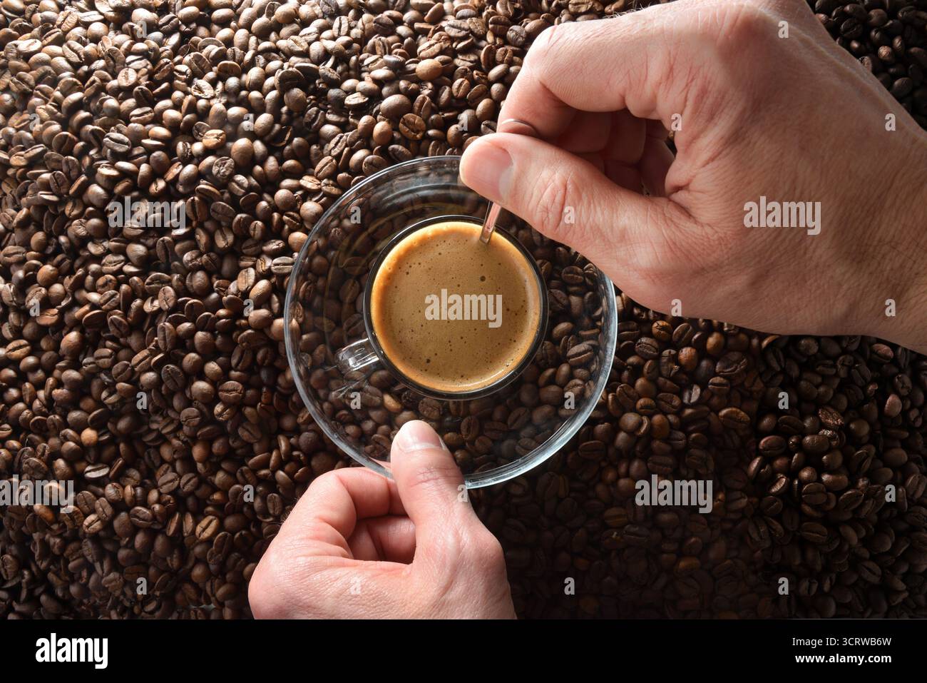 Le mani mescolano il caffè appena estratto in una tazza di vetro e un piattino con un cucchiaio di metallo su una pila di chicchi di caffè. Vista dall'alto. Foto Stock
