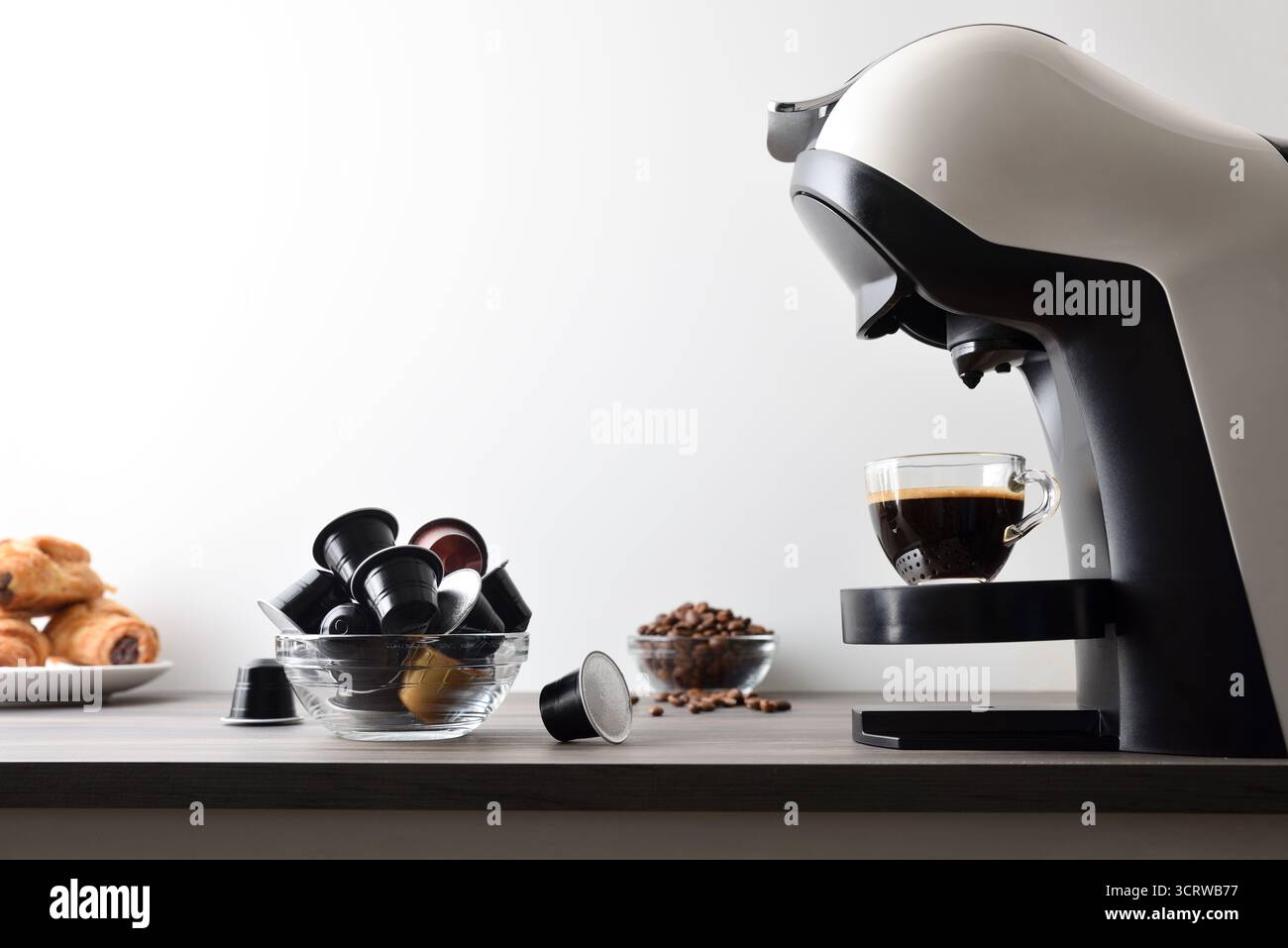 Caffè appena estratto in una macchina espresso su una panca di legno con capsule, chicchi e pasticcini, isolato su sfondo bianco. Vista laterale. Foto Stock
