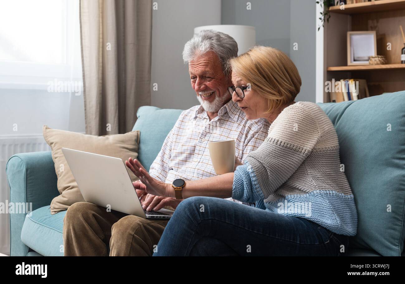 Coppia senior, anziani, vita semplice, seduti a casa sul divano, utilizzando la tecnologia per la prima volta, ordinando e facendo acquisti online su un computer portatile Foto Stock