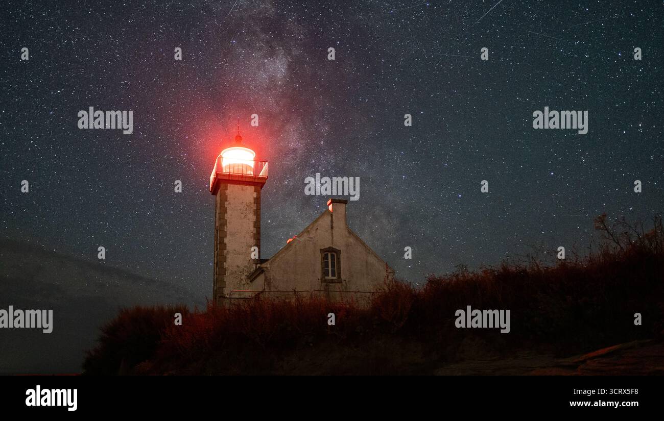 Un faro luminoso si irradia sotto la via Lattea, mescolando il fascino costiero con la magia dell'astrofotografia in un cielo stellato. Foto Stock