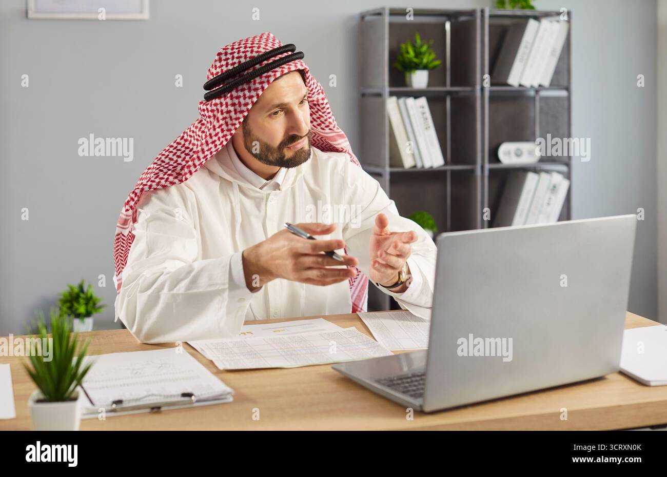 Uomo arabo alla scrivania, indossa abiti tradizionali, guarda lo schermo del notebook, videochiama online Foto Stock