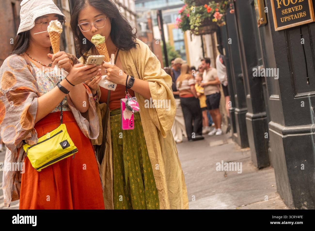 LONDRA - 23 AGOSTO 2025: Turisti asiatici che mangiano gelati e guardano i loro telefoni cellulari a Soho nel West End di Londra Foto Stock
