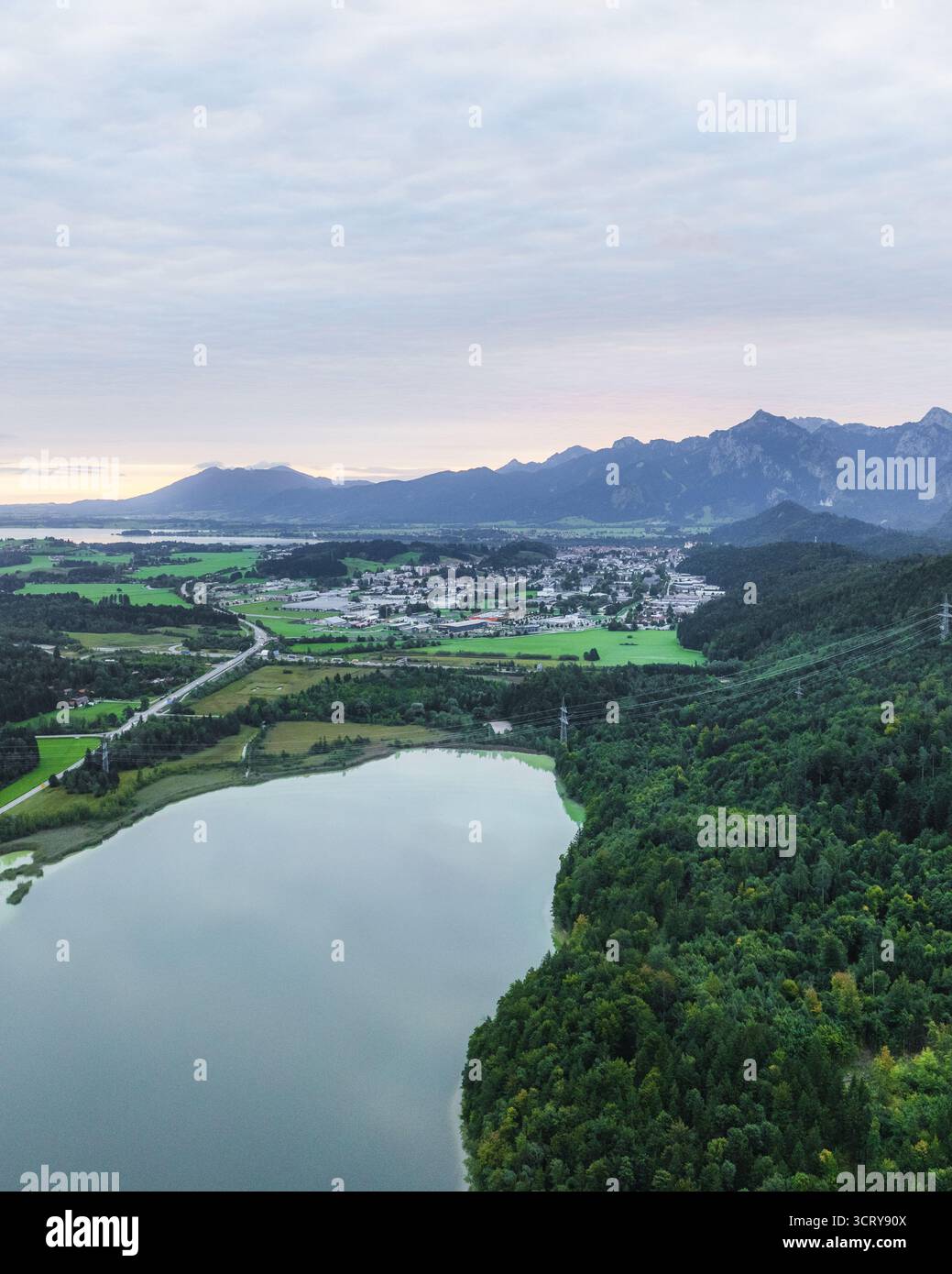 Vista aerea di un placido lago che rispecchia il cielo, delimitato da foreste verdeggianti e incorniciato dalle lontane e maestose Alpi, Füssen, Baviera, Germania. Foto Stock
