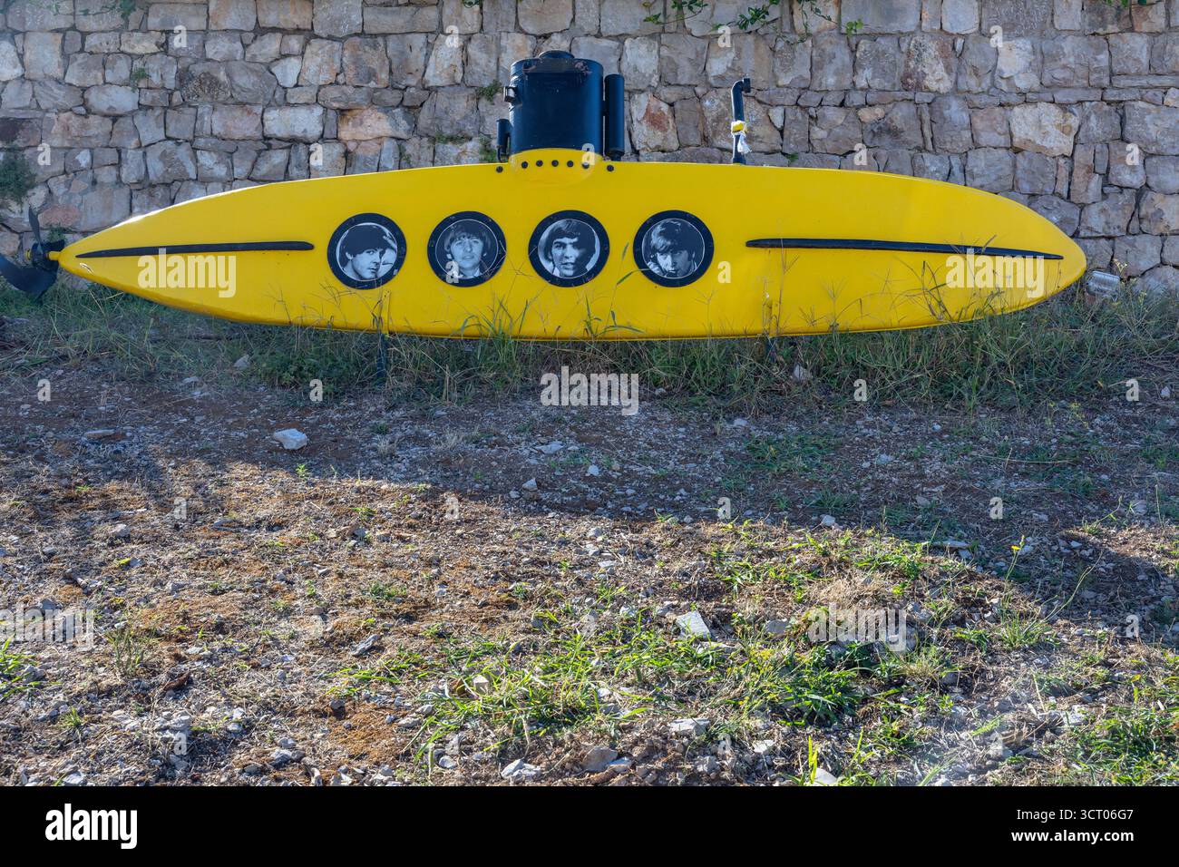MASLINICA, CROAZIA, 08.25.2025: Una scultura subacquea di colore giallo vivo poggia su un prato e roccioso in primo piano contro un grezzo muro di pietra. Quattro oblò circolari Foto Stock