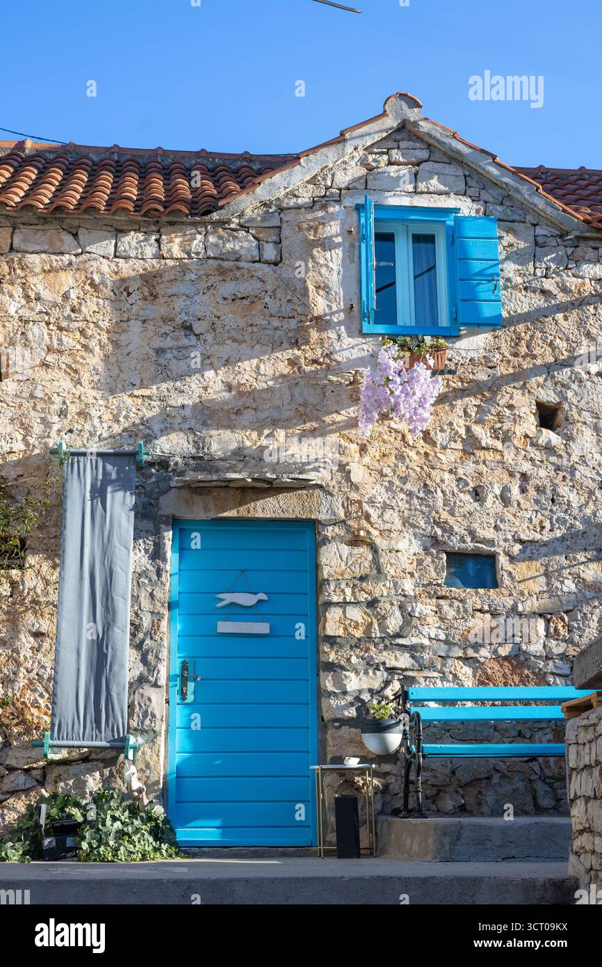 Una casa in pietra intemprata con porte e persiane blu vibranti si trova su una strada mediterranea illuminata dal sole. Ivy si arrampica sul muro, una panchina blu aggiunge fascino, e. Foto Stock