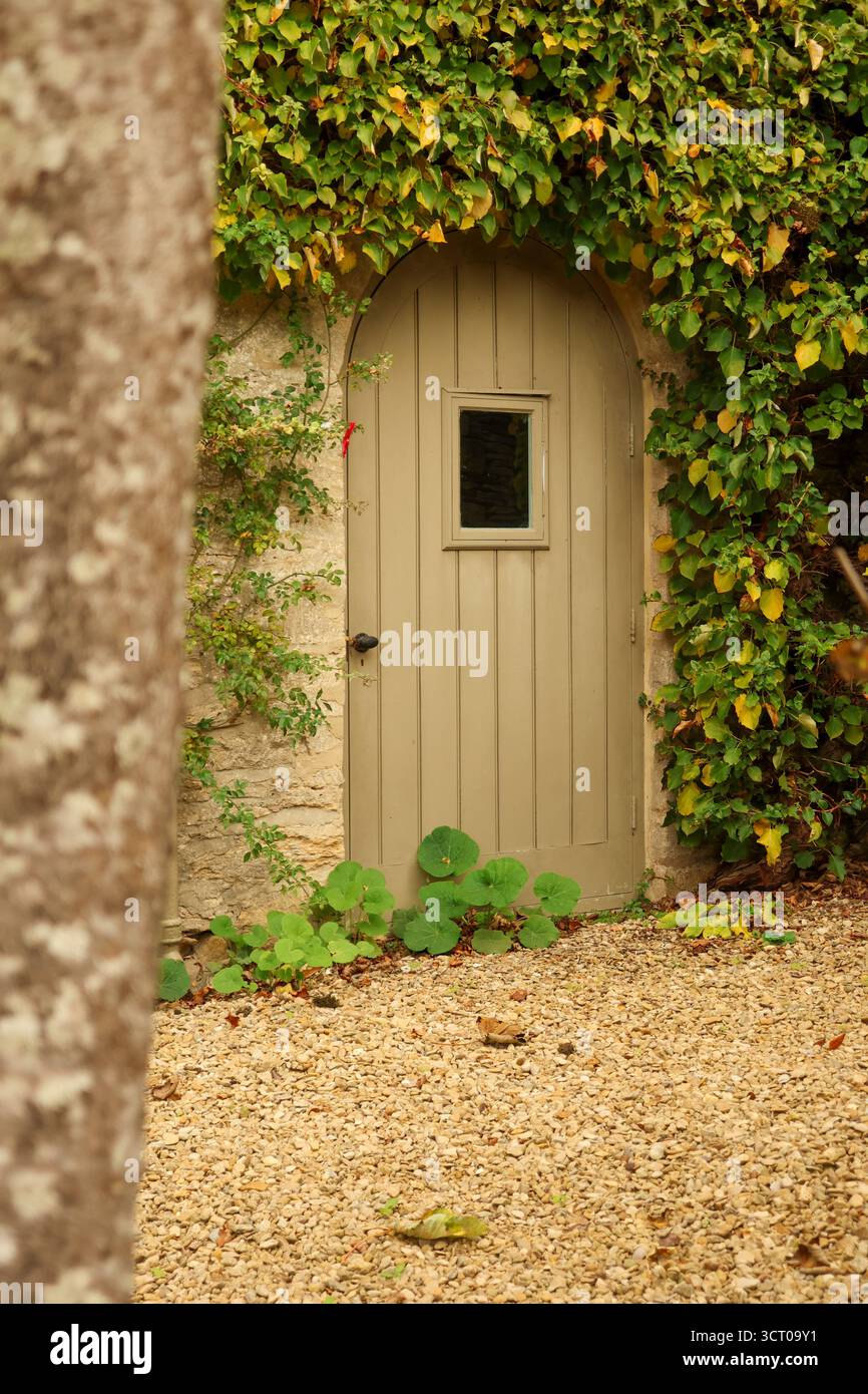 Porta ad arco in legno con finestra incastonata in un muro di pietra, lussureggiante rivestimento in edera, sentiero di ghiaia che conduce all'ingresso segreto del giardino. Cotswolds, Inghilterra Foto Stock