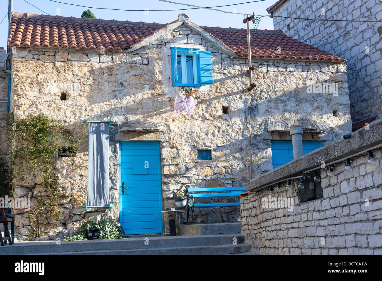 Una casa in pietra intemprata con porte e persiane blu vibranti si trova su una strada mediterranea illuminata dal sole. Ivy si arrampica sul muro, una panchina blu aggiunge fascino, e. Foto Stock