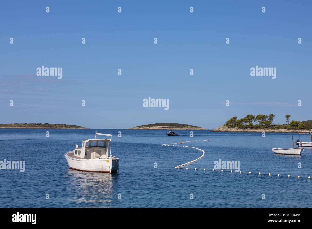 Una tranquilla scena di una barca bianca che galleggia dolcemente su acque blu calme, con una linea curva di boe e isole lontane sotto un cielo luminoso e limpido. Foto Stock