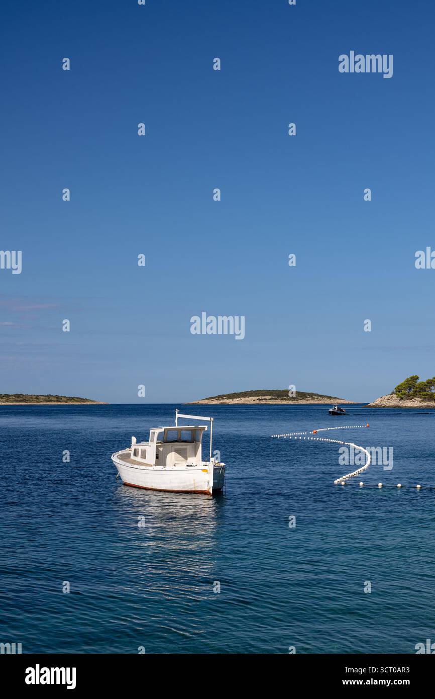Una tranquilla scena di una barca bianca che galleggia dolcemente su acque blu calme, con una linea curva di boe e isole lontane sotto un cielo luminoso e limpido. Foto Stock