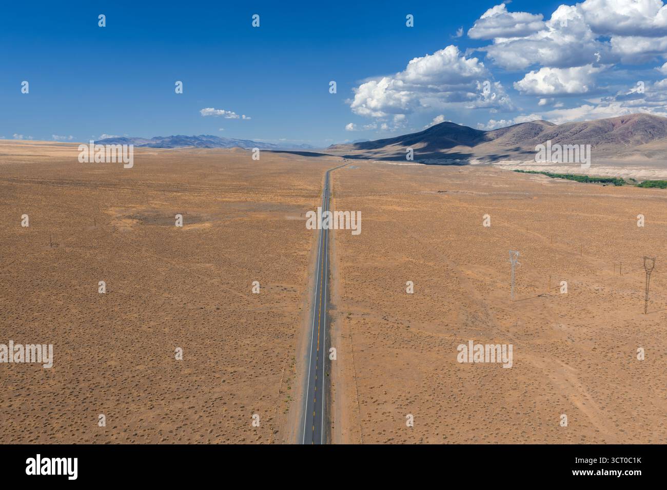 Ampio paesaggio desertico con una strada asfaltata, aspre colline, vegetazione verde, linee elettriche e un cielo blu luminoso con nuvole sparse. Foto Stock