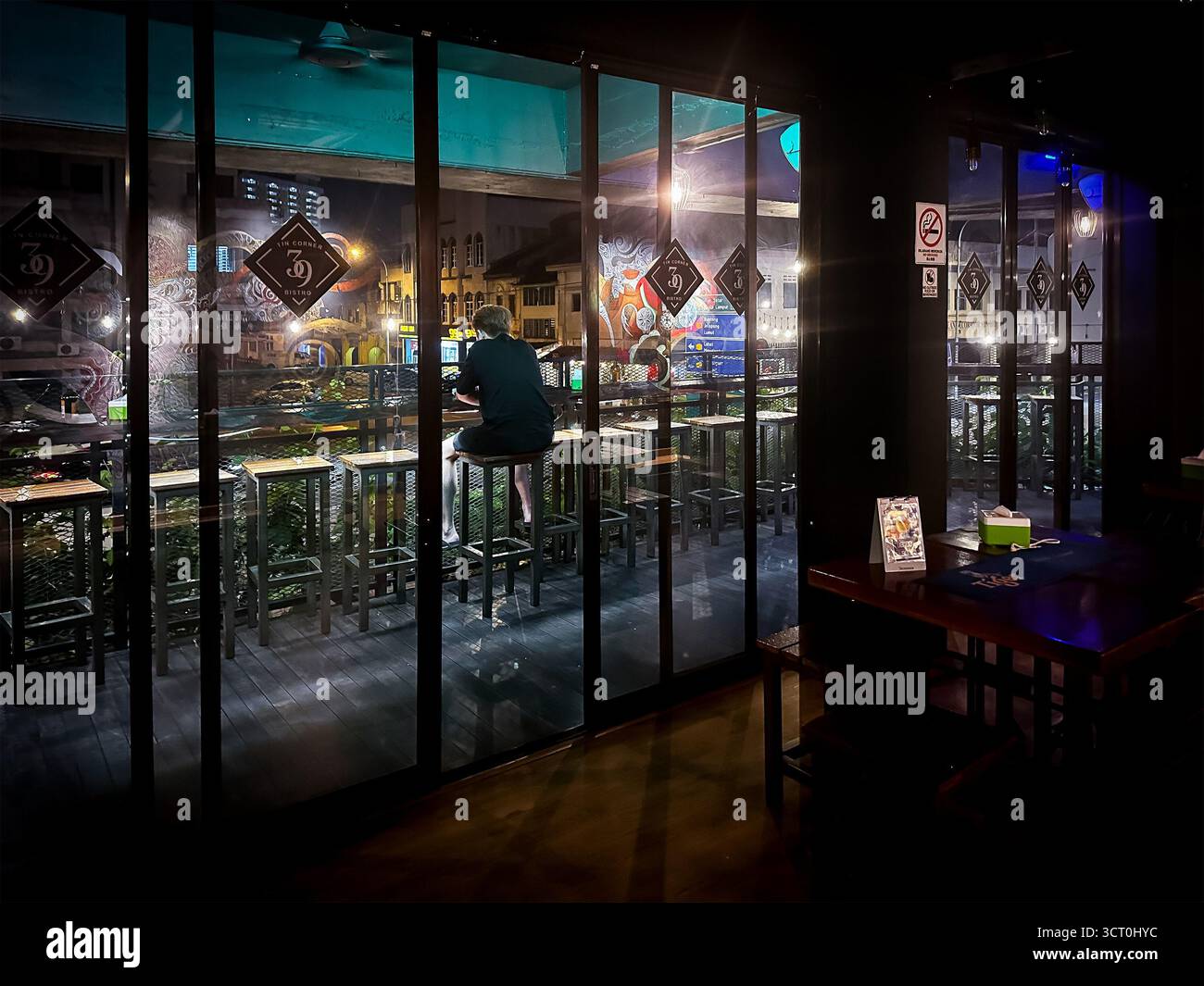 Una figura solitaria siede in un bar in una struttura poco illuminata che si affaccia su una vibrante strada cittadina attraverso grandi finestre di vetro di ipoh malaysia Foto Stock