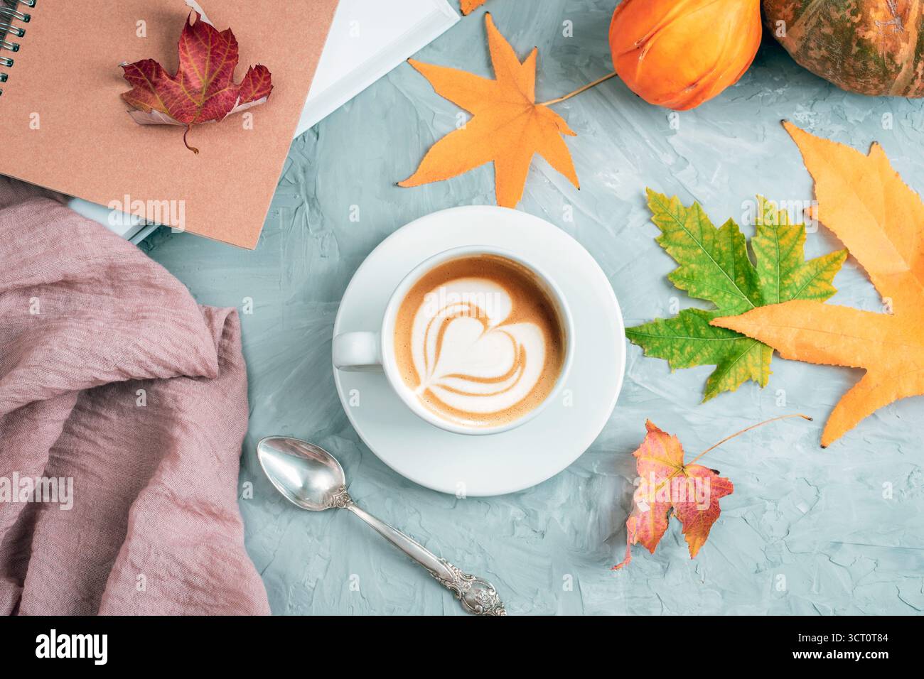 Tazza bianca con cuore di latte art circondata da foglie autunnali, zucche e quaderno su sfondo testurizzato. Concetto autunnale. Vista dall'alto, disposizione piatta. Foto Stock