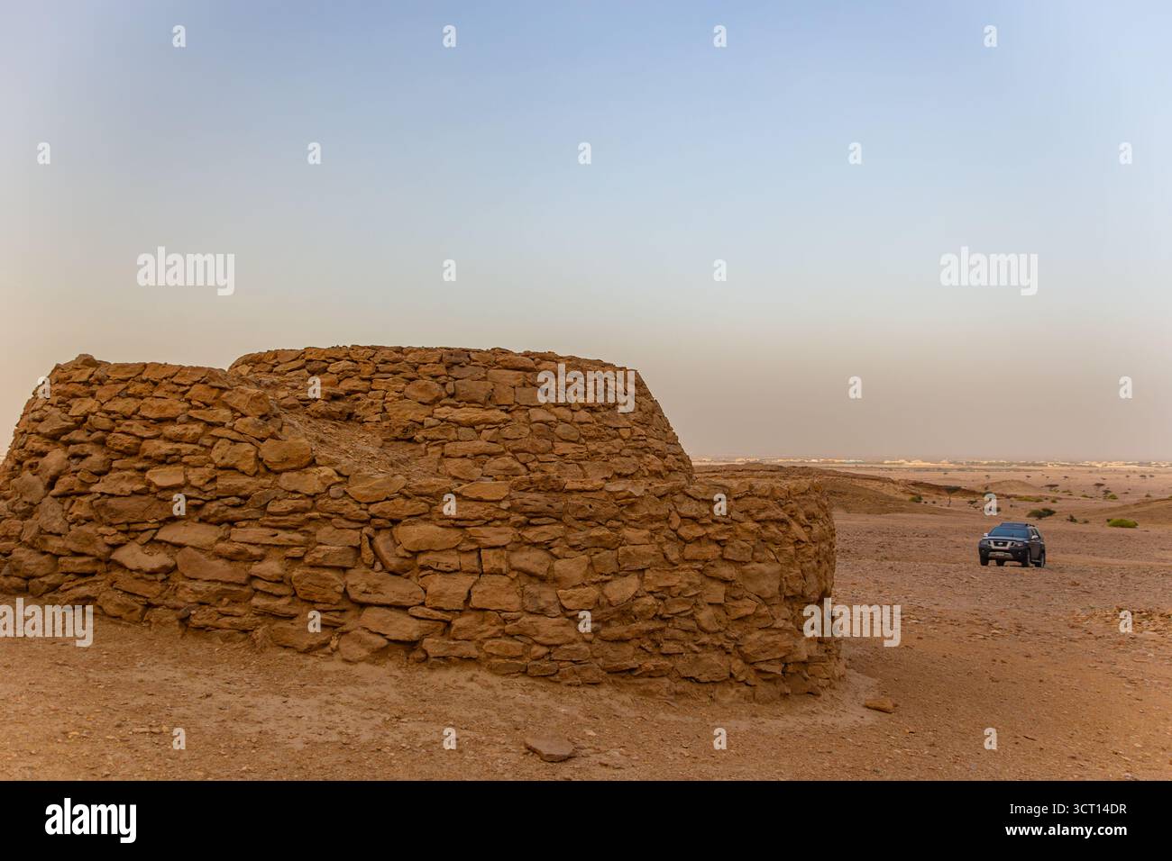 Antica tomba di Beehive realizzata in pietra grezza all'inizio di una vasta e arida pianura del deserto con un moderno SUV parcheggiato in lontananza Foto Stock