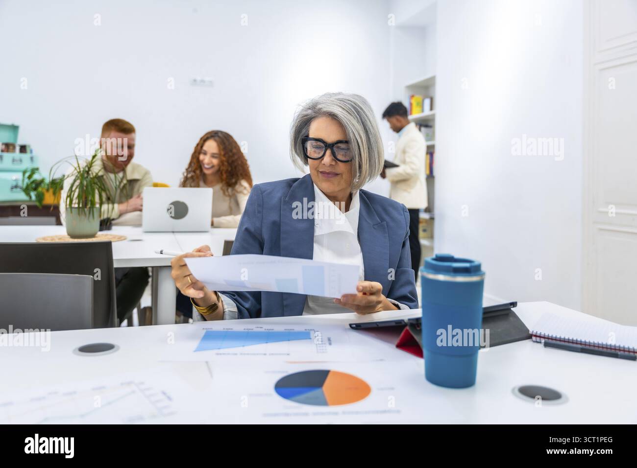 Manager donna matura che esamina un rapporto finanziario con grafici e grafici su un tavolo, collaborando con diversi giovani collaboratori in un ufficio moderno Foto Stock