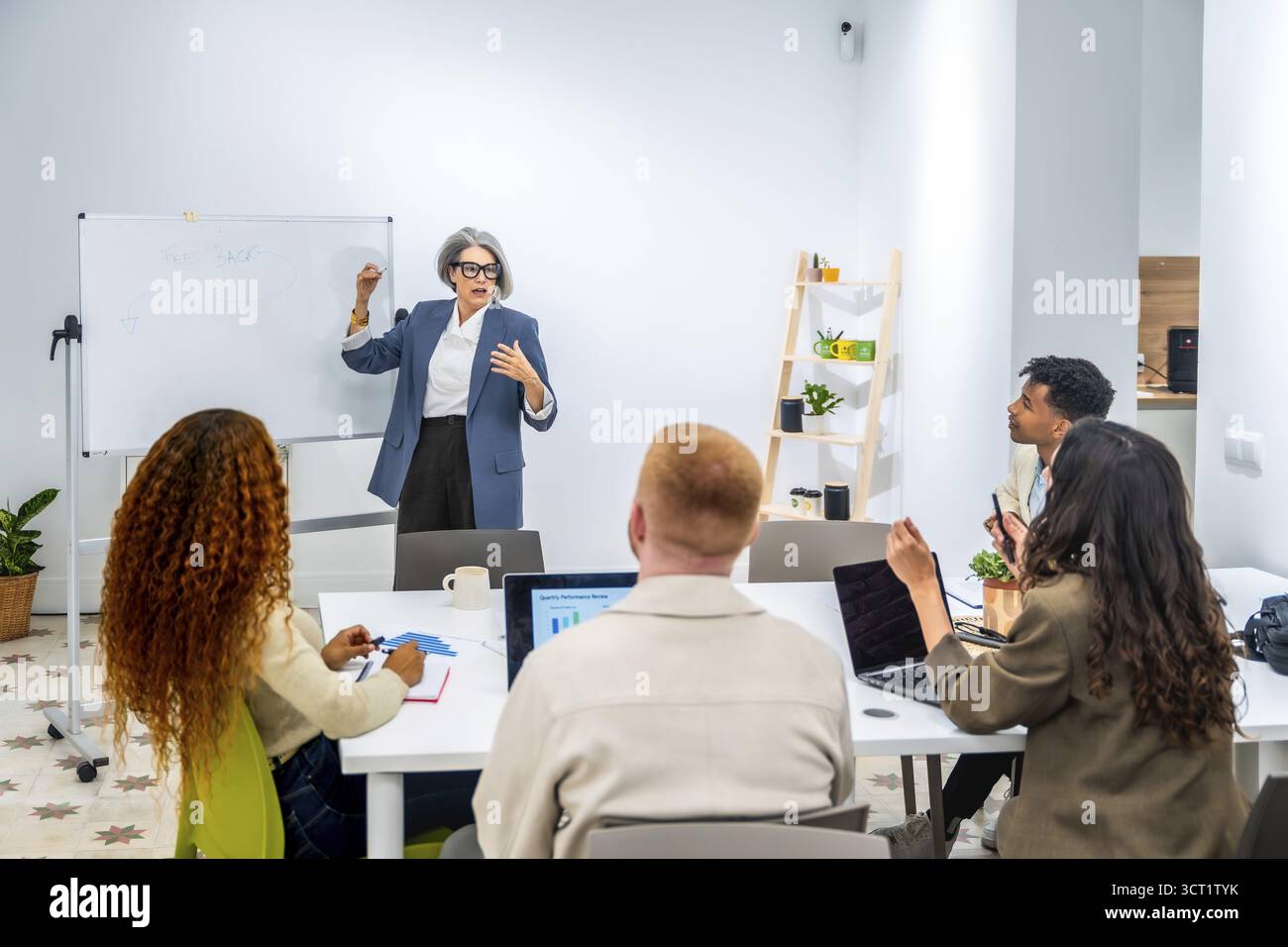 Donna d'affari dai capelli grigi che coaching e formazione un gruppo di giovani professionisti diversi in un ufficio moderno, accanto a una lavagna bianca e all'espla Foto Stock