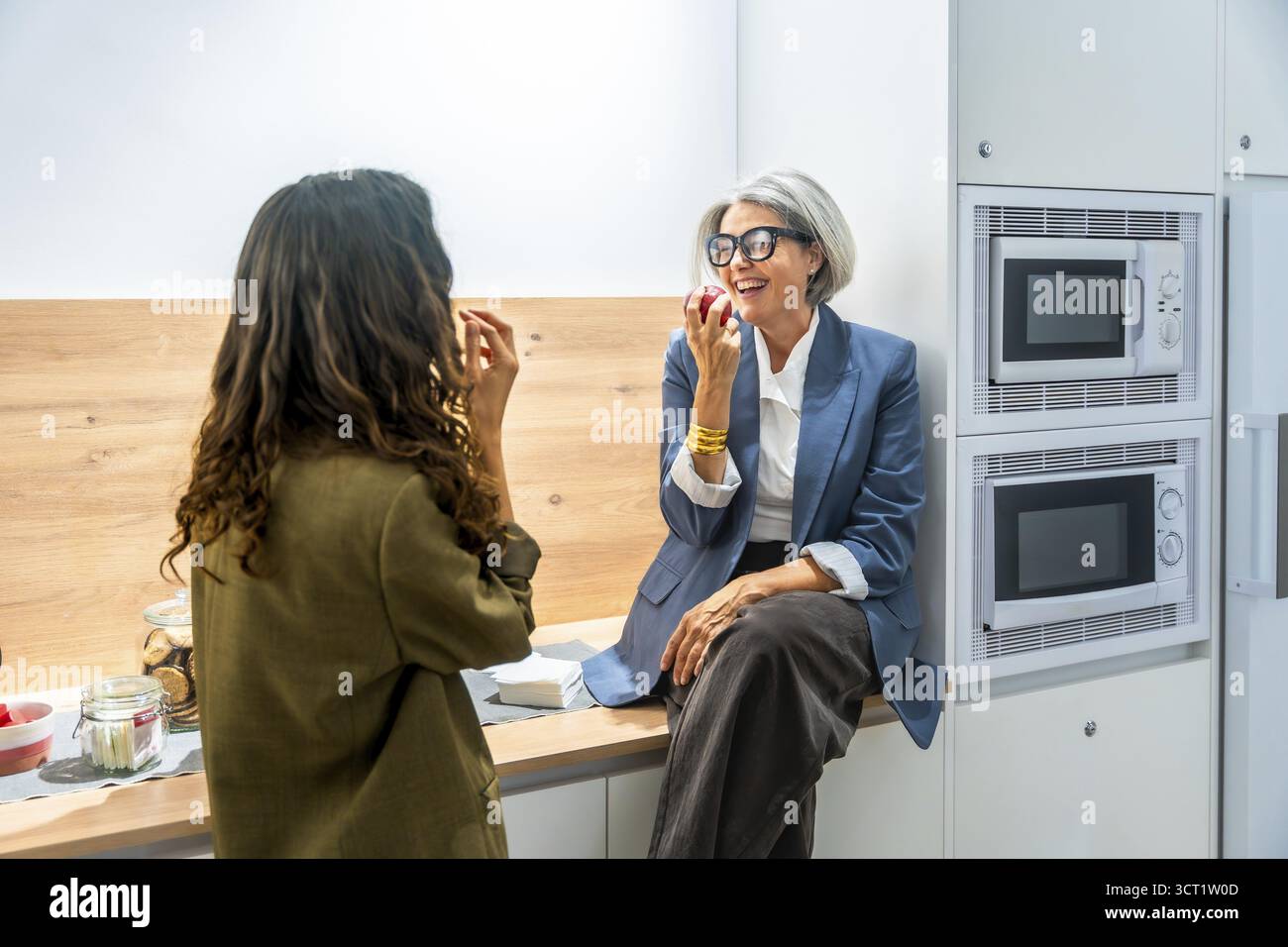 Due donne d'affari colleghi che fanno uno spuntino sano, mangiano mele e ridono insieme mentre fanno una pausa nella moderna cucina dell'ufficio o nella mensa Foto Stock