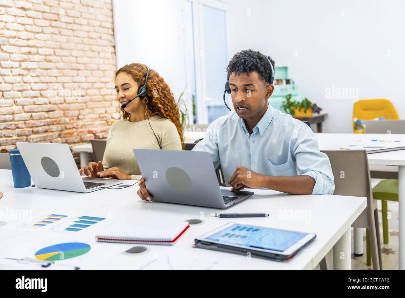 Team di assistenza clienti multirazziale che indossa le cuffie e digita i laptop in un ufficio moderno, fornendo supporto tecnico amichevole, assistenza telefonica e. Foto Stock