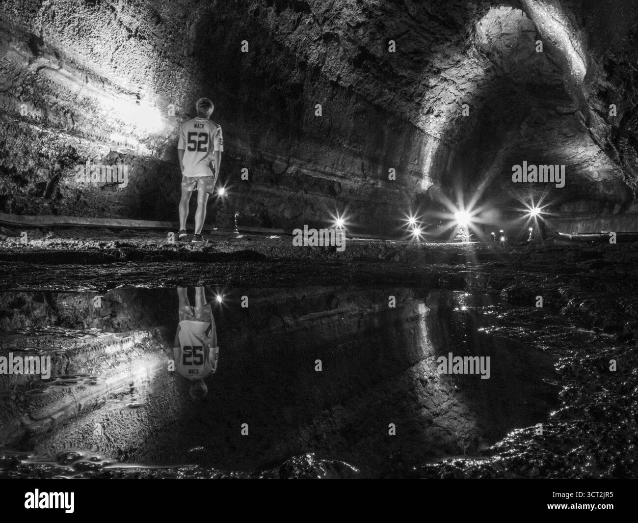 Nella Grotta di Mangjanggul, Jeju, una figura solitaria in una si erge davanti al tunel sul pavimento roccioso accanto a una pozzanghera che riflette la grotta di lava tutta litata Foto Stock