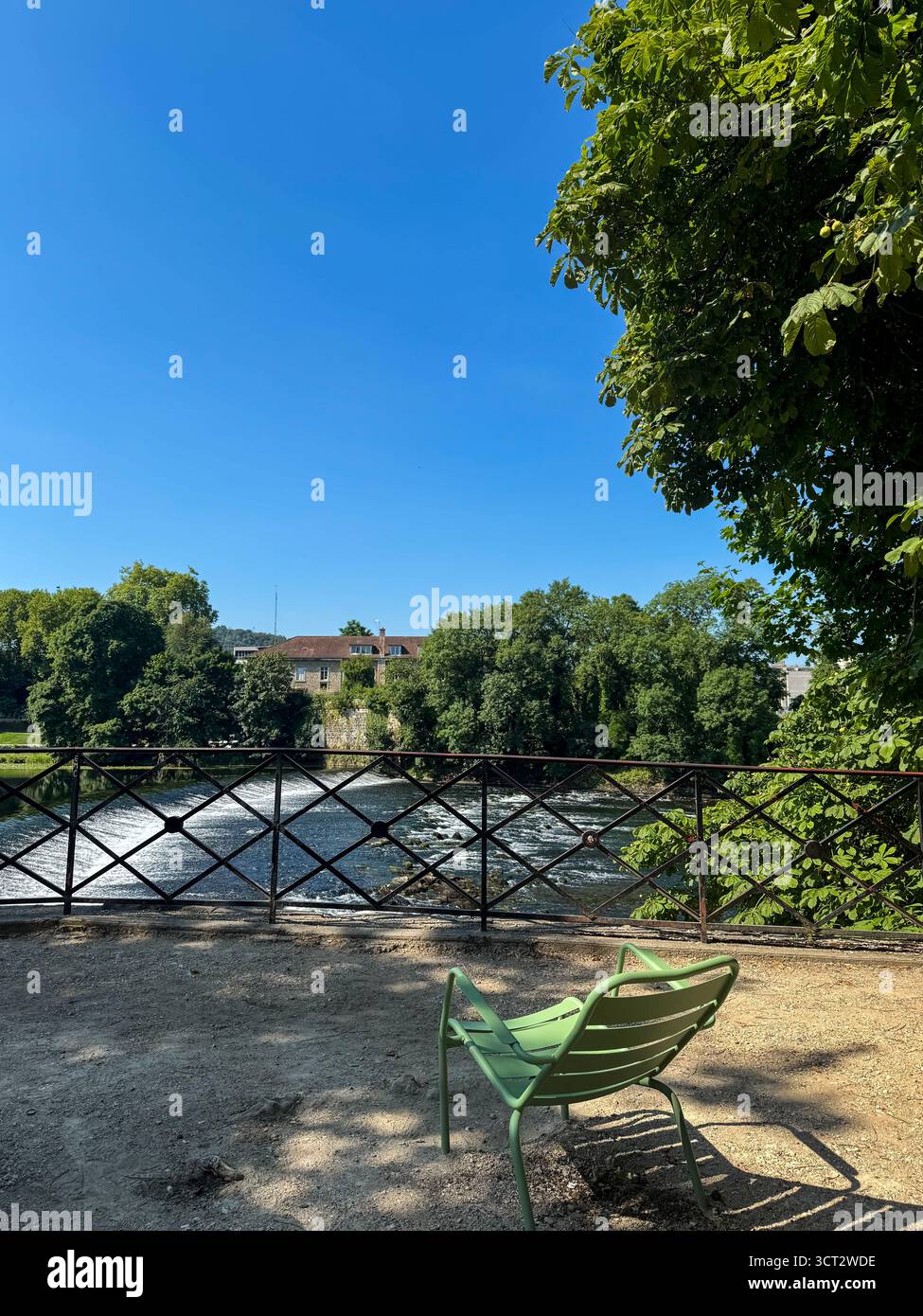Sedia di metallo verde su un sentiero di ghiaia che si affaccia sul fiume Doubs in un tranquillo parco cittadino sotto un cielo azzurro. - Immagine stock catturata con smartphone