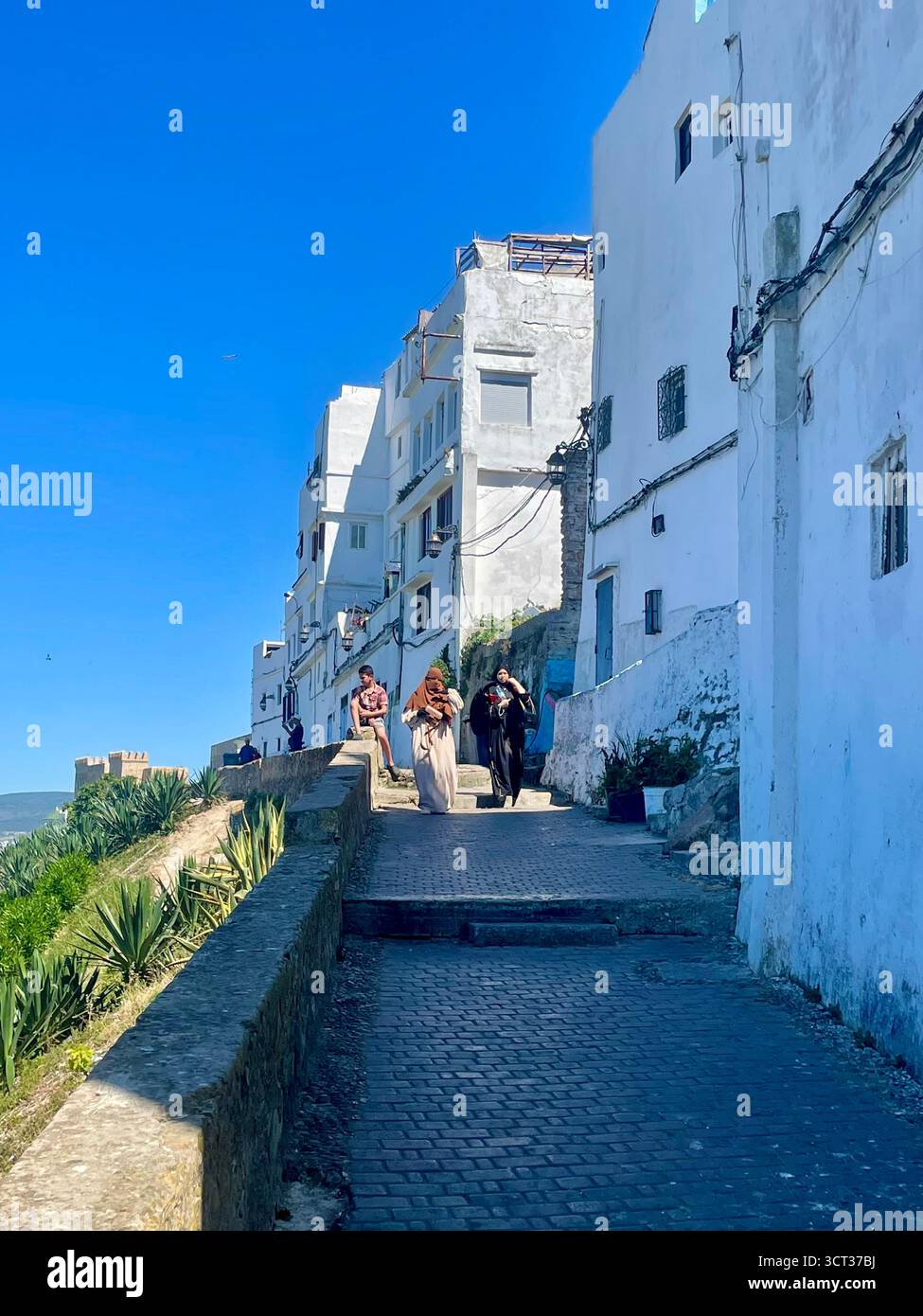 Sentiero acciottolato con edifici bianchi e figure tradizionali sotto un cielo limpido. Tangeri, Marocco — 7 giugno 2024. Foto Stock