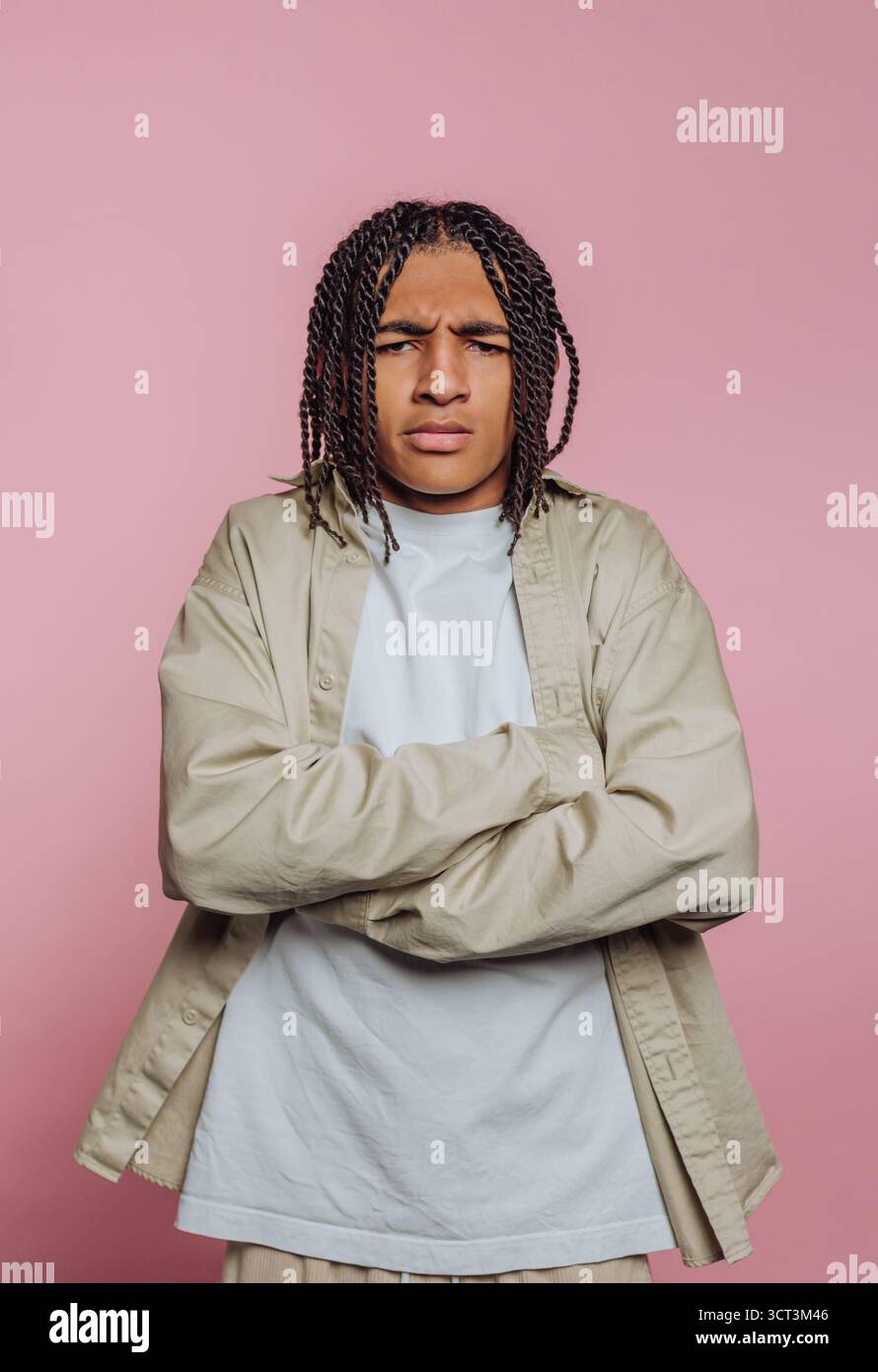 Un giovane uomo sta con le braccia incrociate, mostrando un'espressione seria. È vestito in modo informale con una camicia e una giacca di colore chiaro, con un dorso rosa Foto Stock
