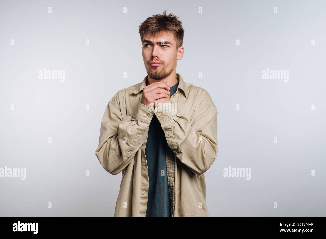 Un giovane si trova in un semplice studio, indossando una camicia beige chiara sopra una camicia scura. Sembra pensivo, con le mani legate insieme. Il backgroun Foto Stock