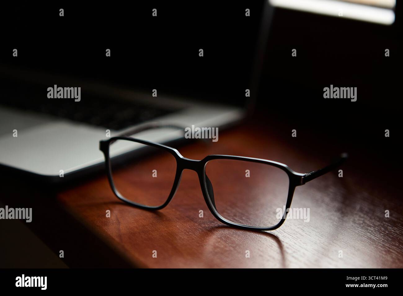 Un paio di occhiali da vista posizionati su una scrivania in legno lucidato accanto a un laptop aperto, per creare un ambiente di lavoro elegante con un'atmosfera professionale Foto Stock