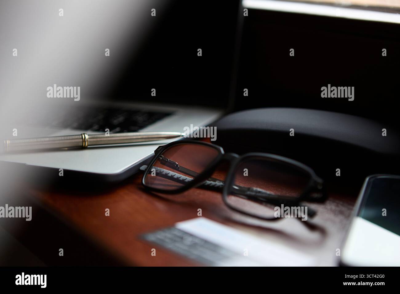 Una vista ravvicinata degli occhiali da vista che si trovano accanto a una penna e a un computer portatile su una scrivania in legno lucido con riflessi morbidi. L'immagine mostra un lavoro minimalista ordinato Foto Stock