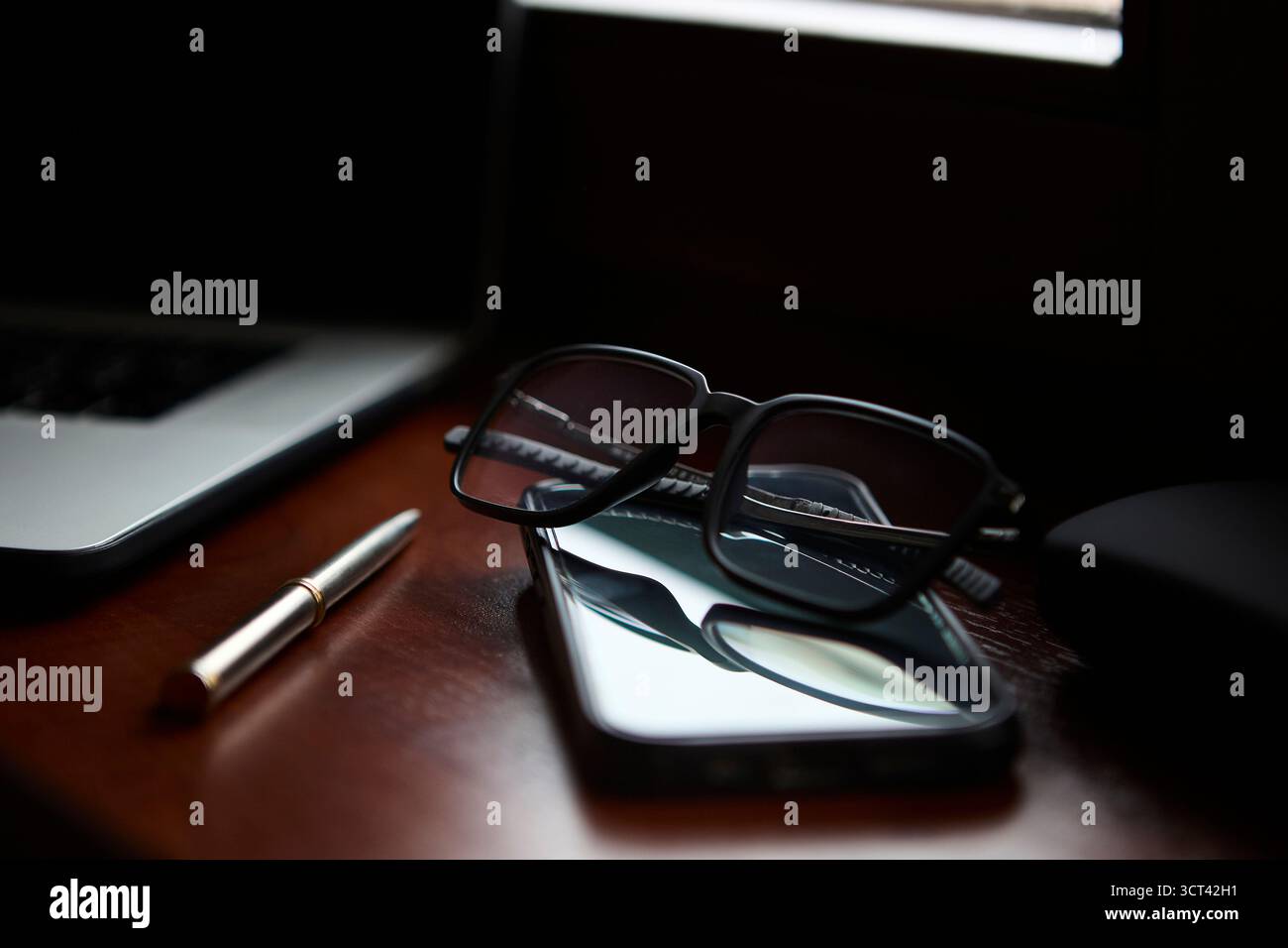 Un elegante paio di occhiali da vista si trova sopra uno smartphone con una penna nelle vicinanze su una scrivania in legno lucido. Il notebook sfocato sullo sfondo lo consiglia Foto Stock