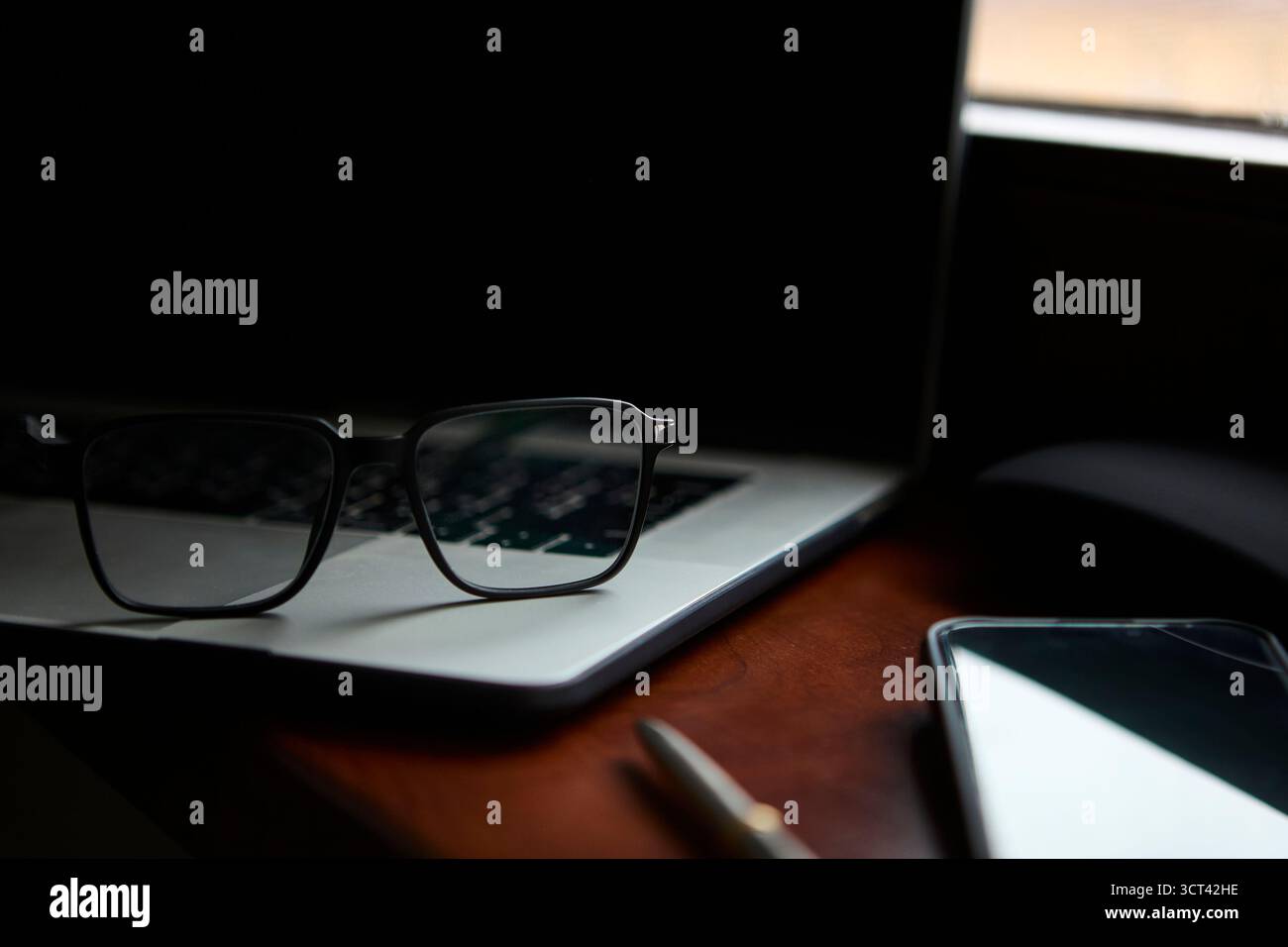 Gli occhiali sono posizionati sulla tastiera di un computer portatile con una penna e uno smartphone posizionati sulla scrivania accanto. L'ambiente trasmette un moderno business o accademico Foto Stock