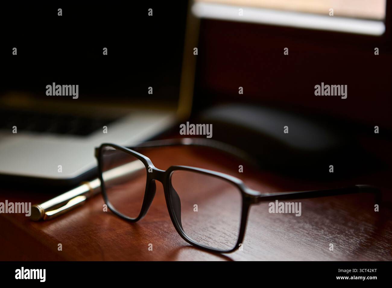 Un primo piano di occhiali da lettura appoggiati su una scrivania in legno lucido accanto a una penna dorata e a un computer portatile. La composizione crea un ufficio professionale elegante Foto Stock