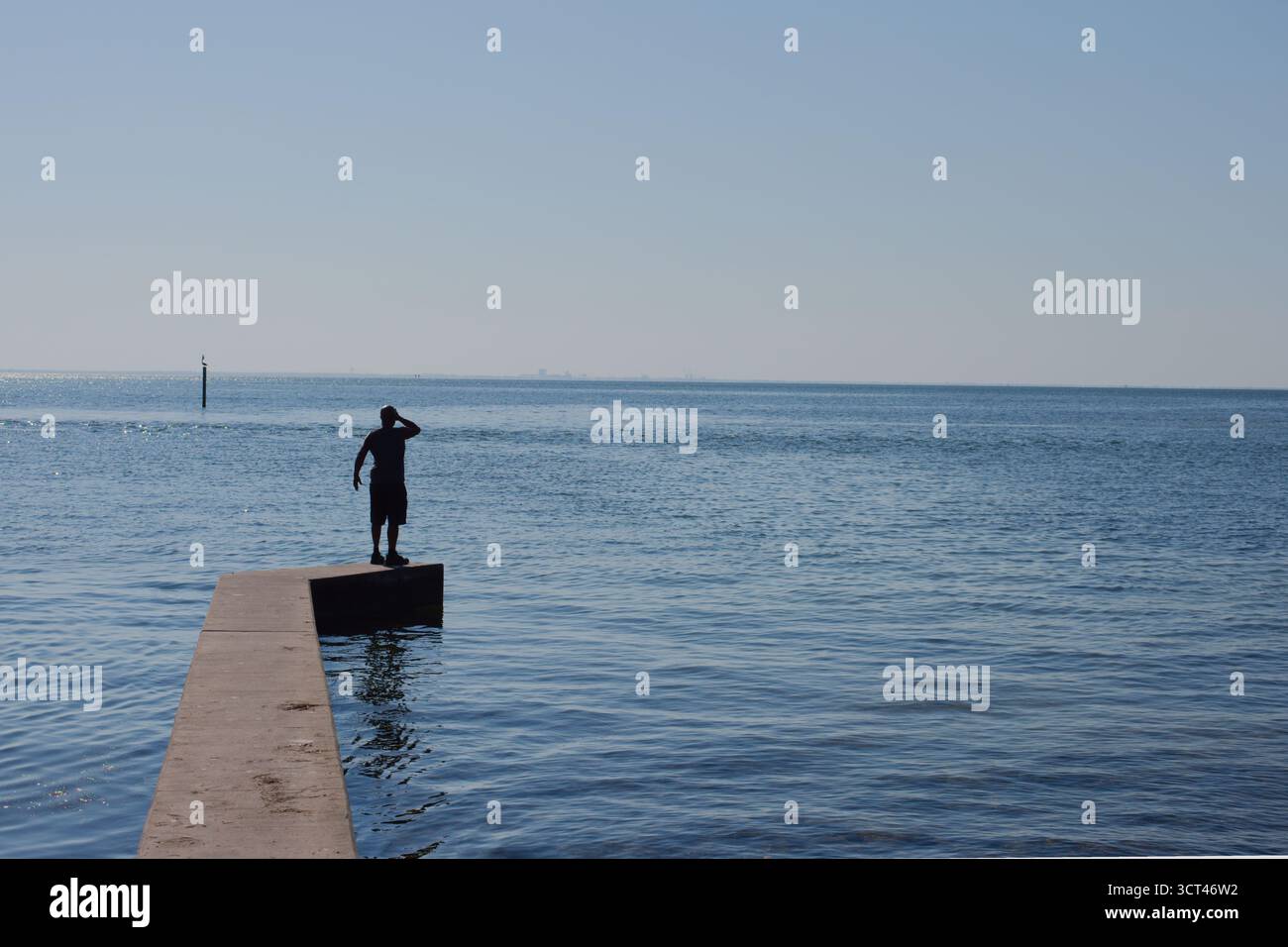 Silhouette di un uomo solitario su uno stretto molo che guarda il mare aperto sotto il cielo azzurro a St Petersburg, Florida, vista sulla baia di Tampa. Fine di un calcestruzzo Foto Stock