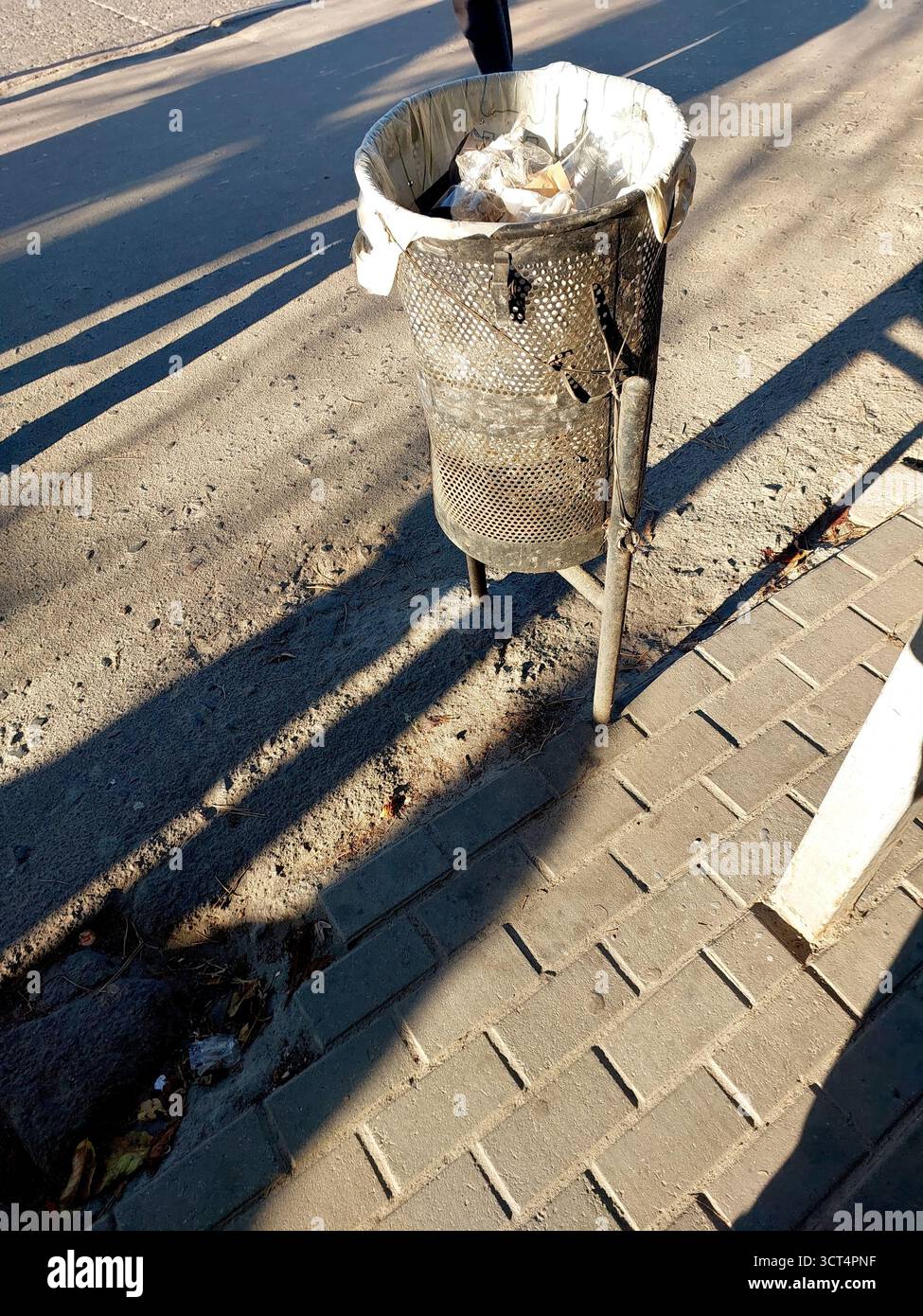 Un cestino a rete nera si trova su una strada con la luce del sole che proietta lunghe ombre. Contiene articoli scartati e si trova vicino a un marciapiede. Foto Stock