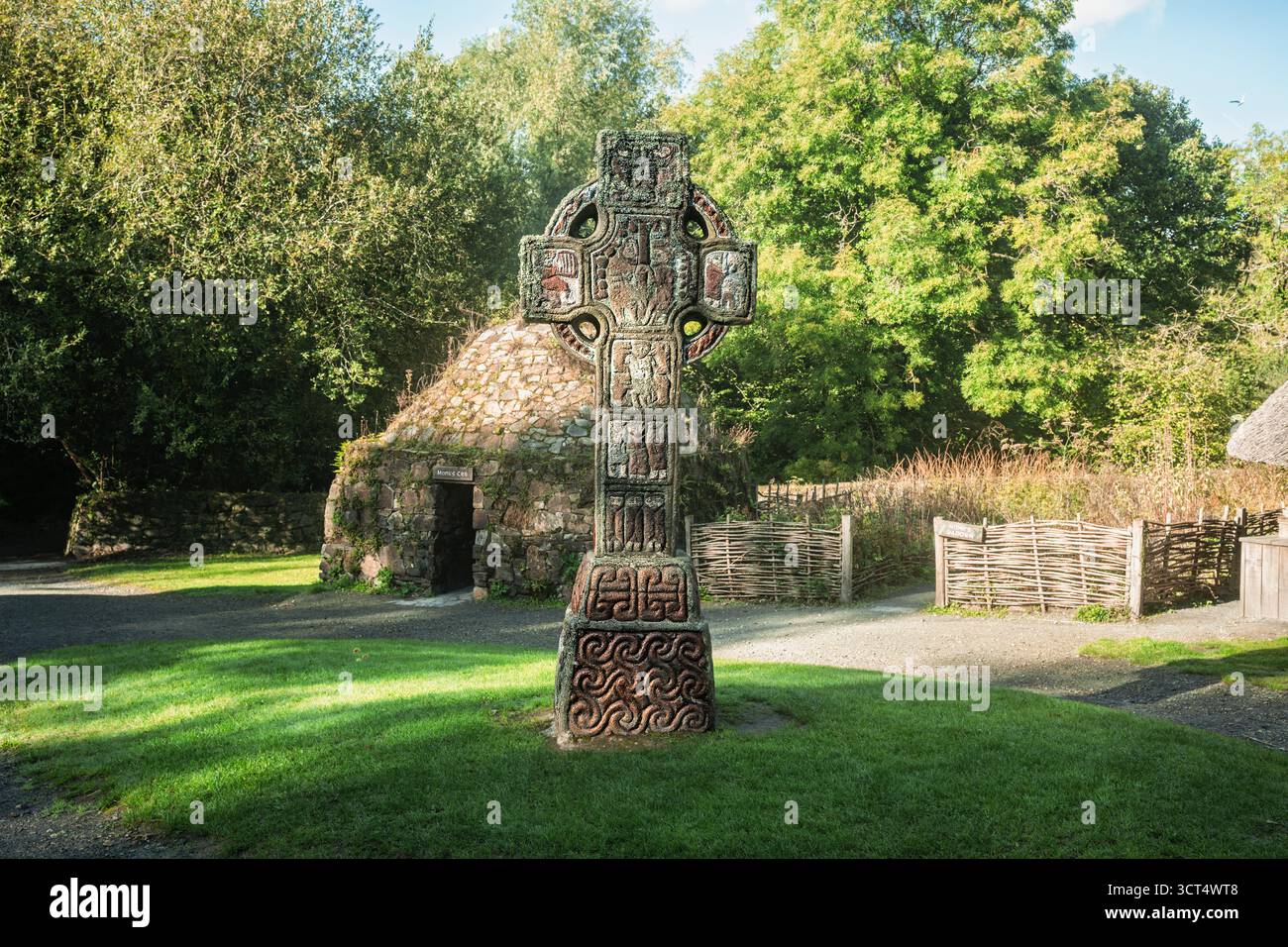 Alta Croce paleocristiana con pannelli scolpiti accanto alla pietra clochán all'Irish National Heritage Park Foto Stock