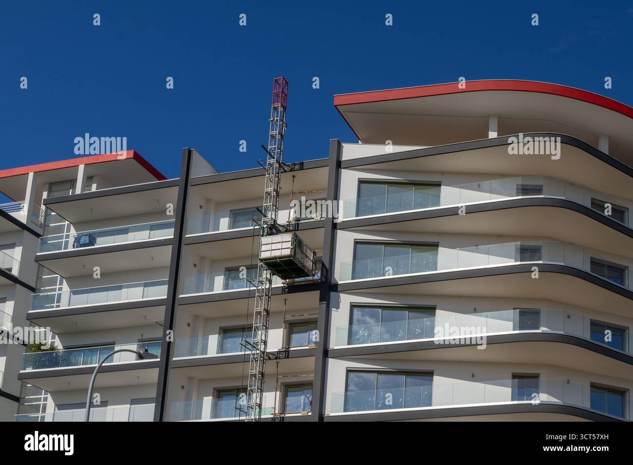 Costruzione di ascensore che solleva i materiali sulla moderna facciata dell'edificio con balconi in vetro sotto il cielo blu Foto Stock
