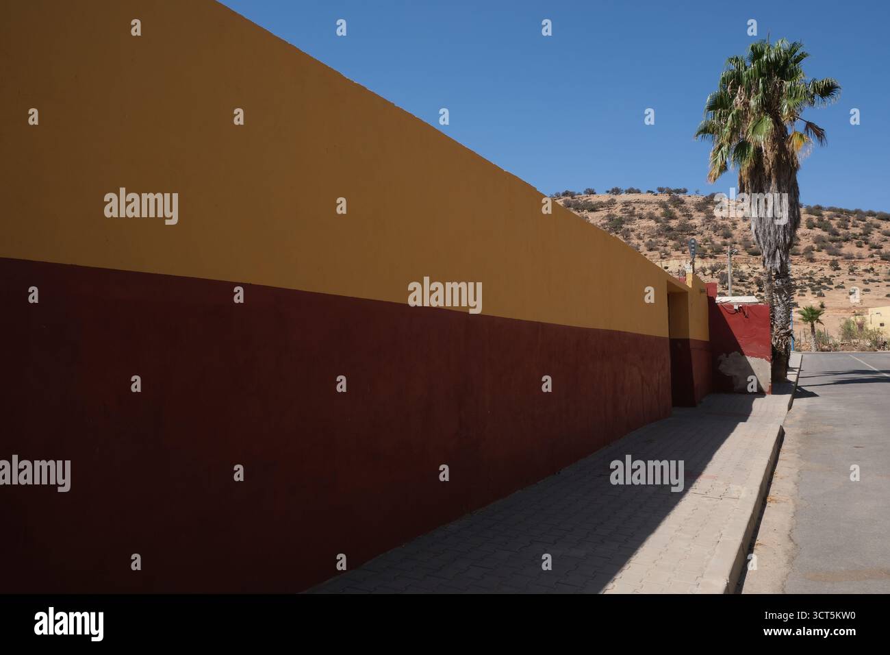 Una parete dipinta di giallo e rosso brillante che termina con una palma, una strada nella campagna del Marocco del Nord Africa Foto Stock