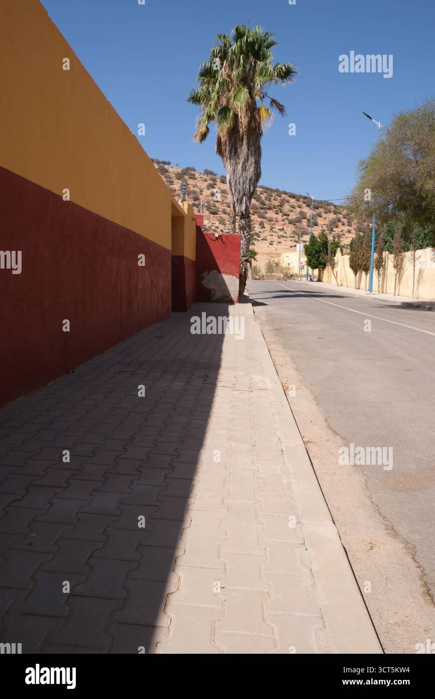 Una parete dipinta di giallo e rosso brillante che termina con una palma, una strada nella campagna del Marocco del Nord Africa Foto Stock