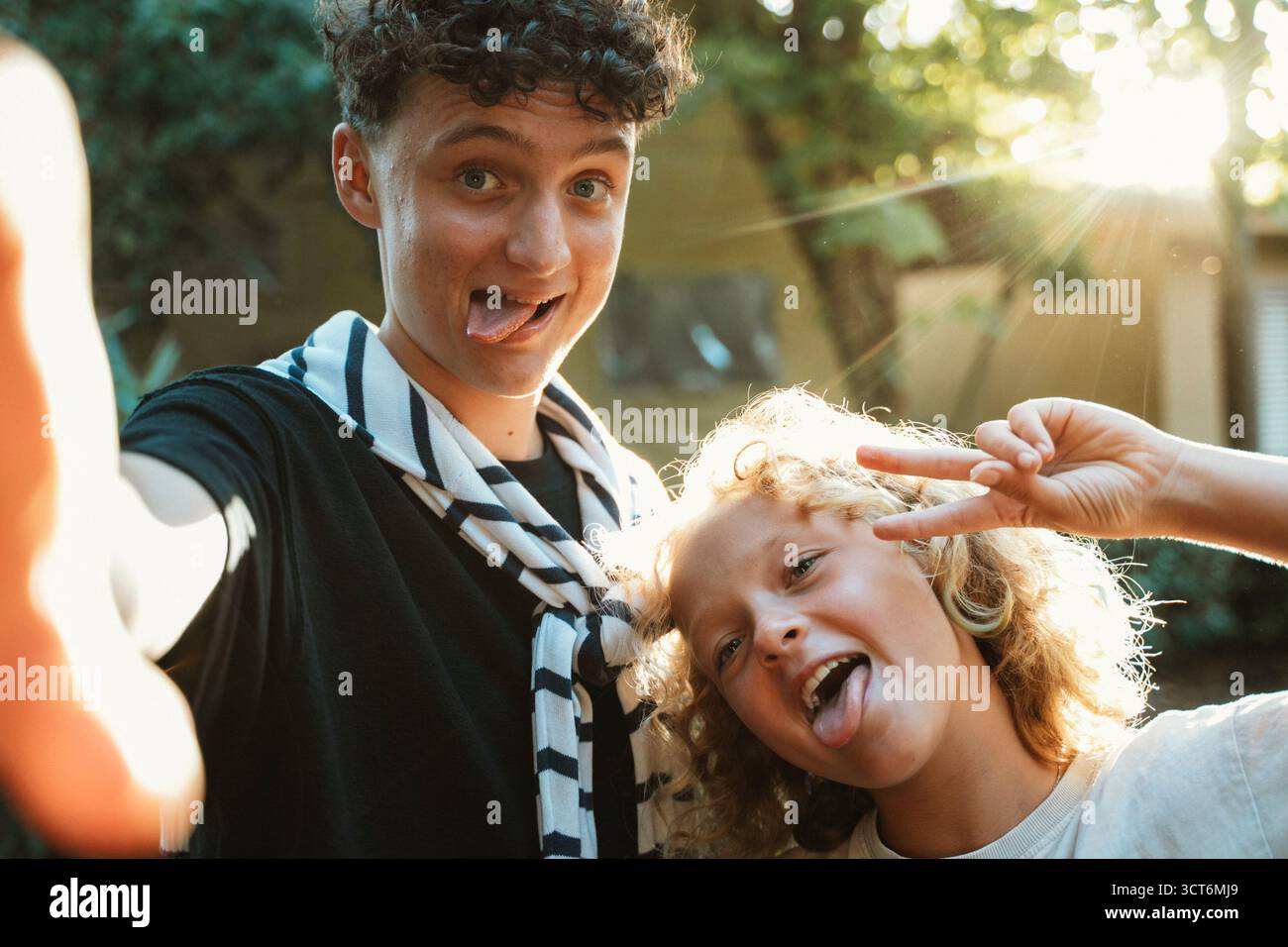Fratello e sorella esprimono gioia con facce divertenti, mostrando gesti giocosi della lingua e segni di pace. Catturati in un ambiente all'aperto illuminato dal sole, che riflette le labbra Foto Stock