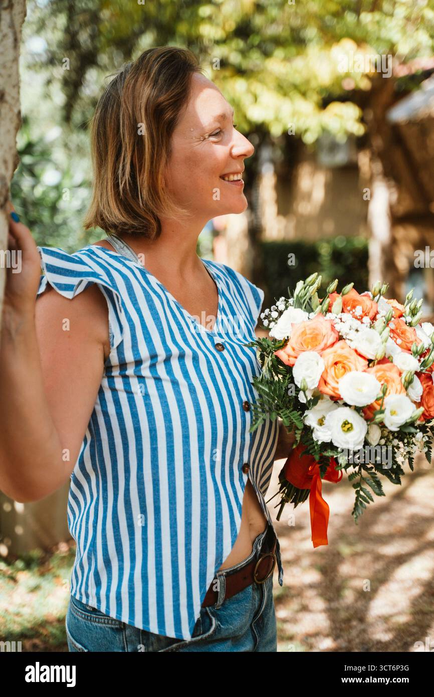 Una donna che indossa una maglia a righe sorride mentre tiene in mano un vivace bouquet di fiori in un ambiente all'aperto. La sua espressione gioiosa e la composizione floreale crea Foto Stock