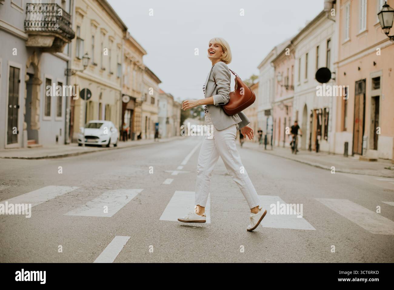 Una donna con i capelli corti passeggia lungo un incrocio in una tranquilla zona urbana. Indossa abiti casual e porta con sé una borsa, trascorrendo una piacevole giornata Foto Stock