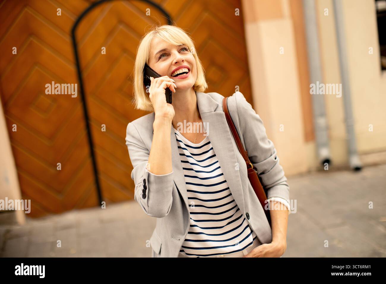 Una giovane donna con i capelli biondi è impegnata in una conversazione telefonica allegra. Indossa un elegante blazer grigio su una camicia a righe e si distingue per il suo fascino Foto Stock