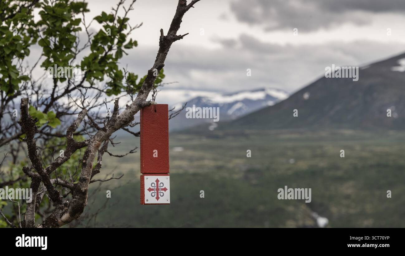 Wegmarkierung in forma eines rot-weissen Schildes haengt an einem AST, Olavskreuz fuer Pilger, im Hintergrund Berge und Wolken, Olavsweg, Olavsleden od Foto Stock