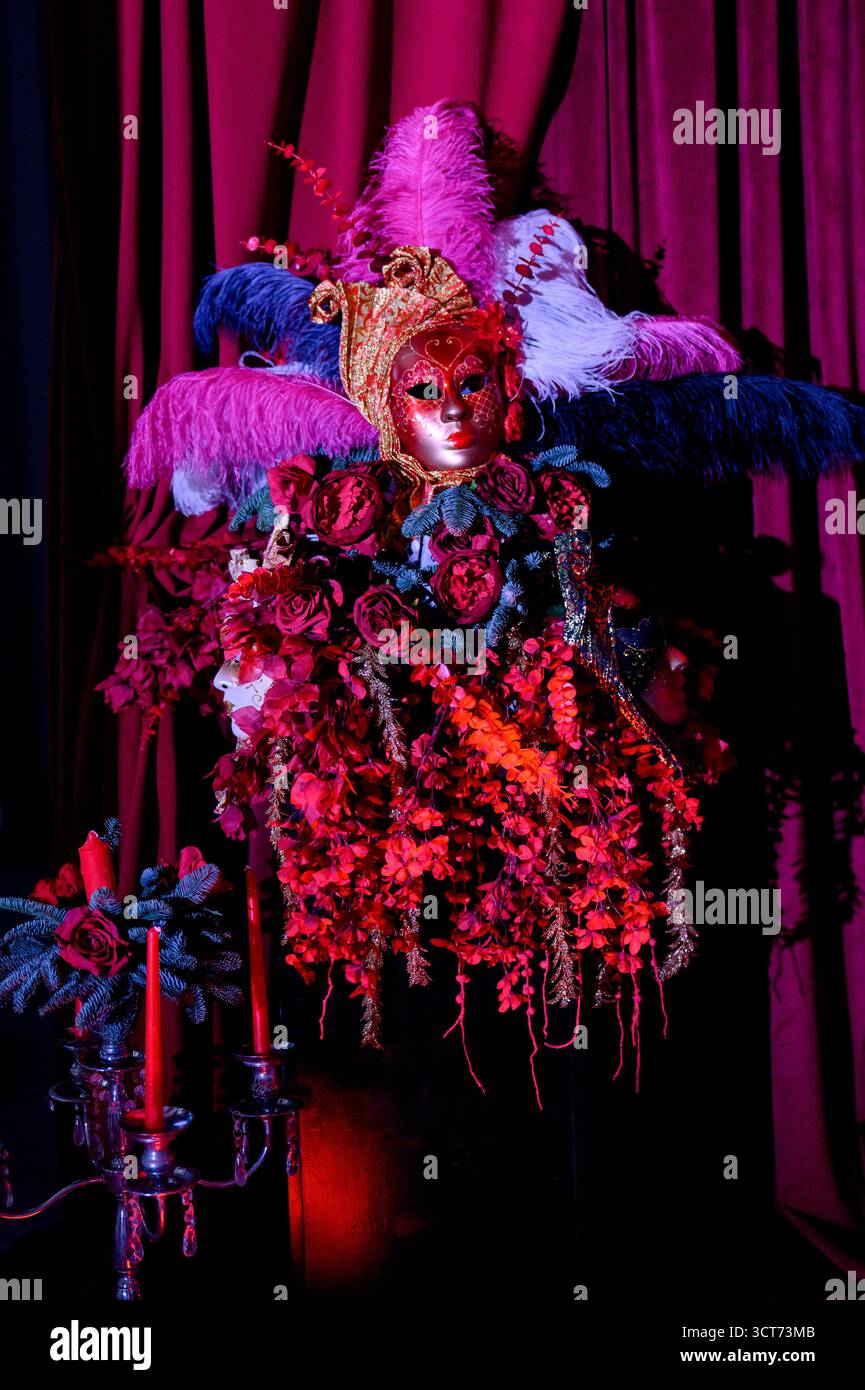 Decorazioni per feste con maschere, candele e piume sullo sfondo di profonde tende di velluto viola. Elementi decorativi del Carnevale con maschere veneziane in rosso e. Foto Stock