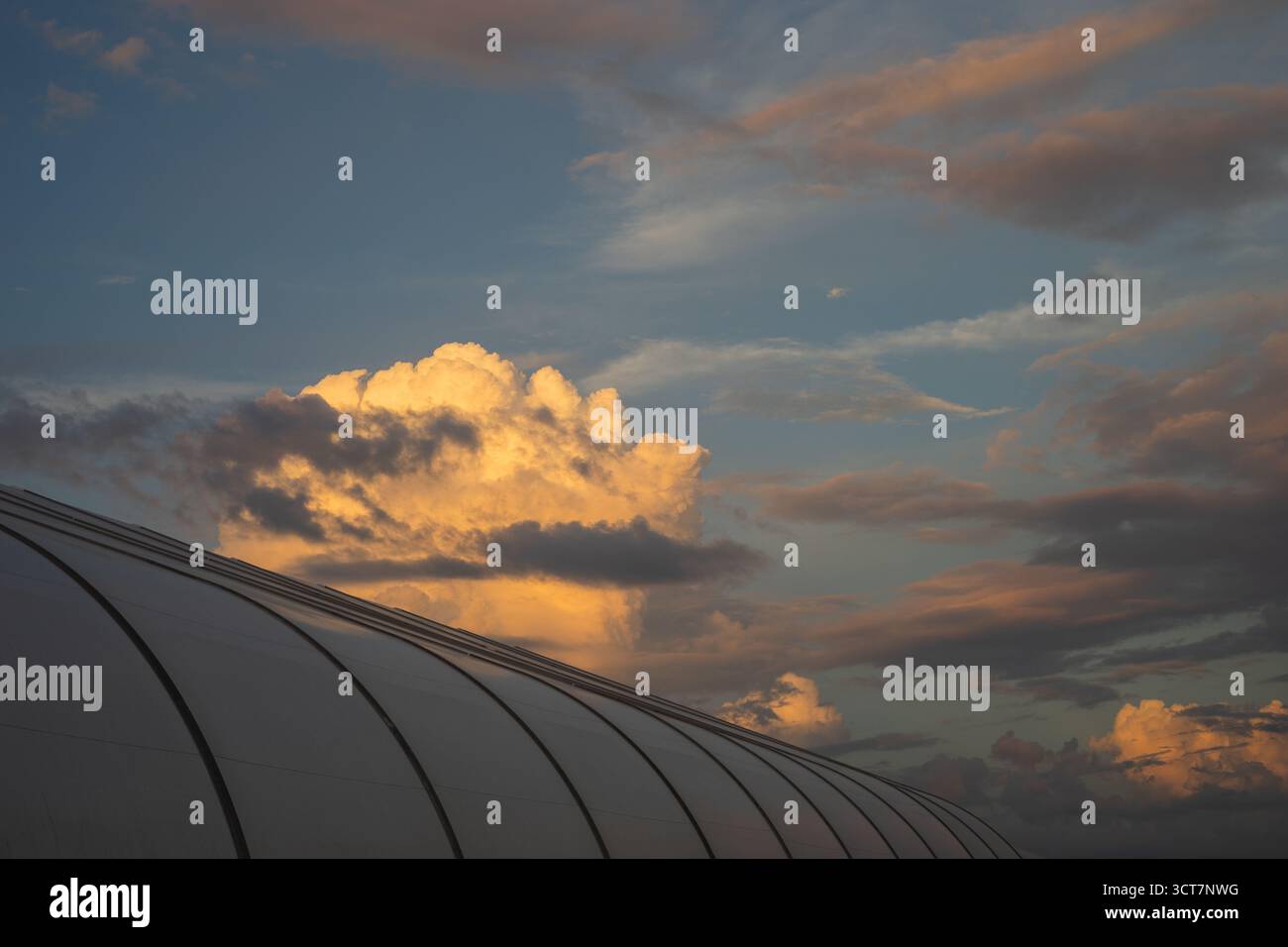 Nuvole dorate al tramonto sopra una moderna struttura a tetto ricurvo, creando un contrasto tra architettura e natura nella calda luce serale. Foto Stock