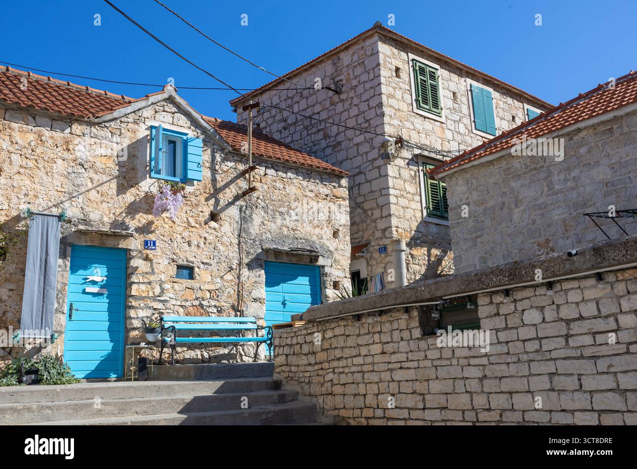 Una casa in pietra intemprata con porte e persiane blu vibranti si trova su una strada mediterranea illuminata dal sole. Ivy si arrampica sul muro, una panchina blu aggiunge fascino, e. Foto Stock