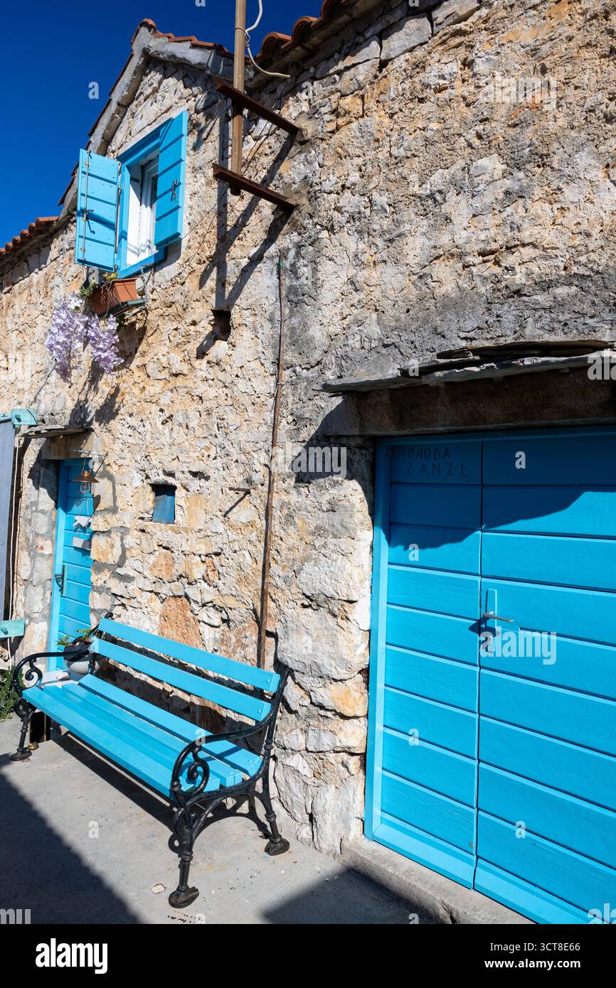Una casa in pietra intemprata con porte e persiane blu vibranti si trova su una strada mediterranea illuminata dal sole. Ivy si arrampica sul muro, una panchina blu aggiunge fascino, e. Foto Stock